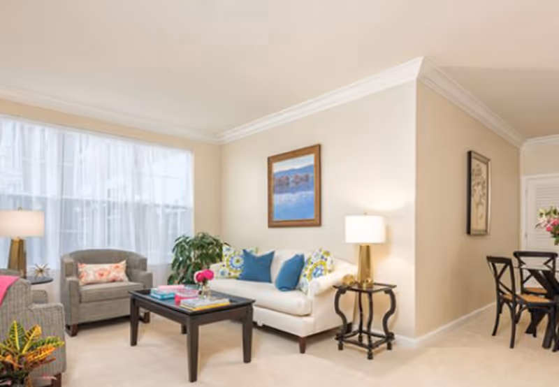 Bright living room with a white sofa, armchairs, coffee table, lamps, and a dining area visible in the background.