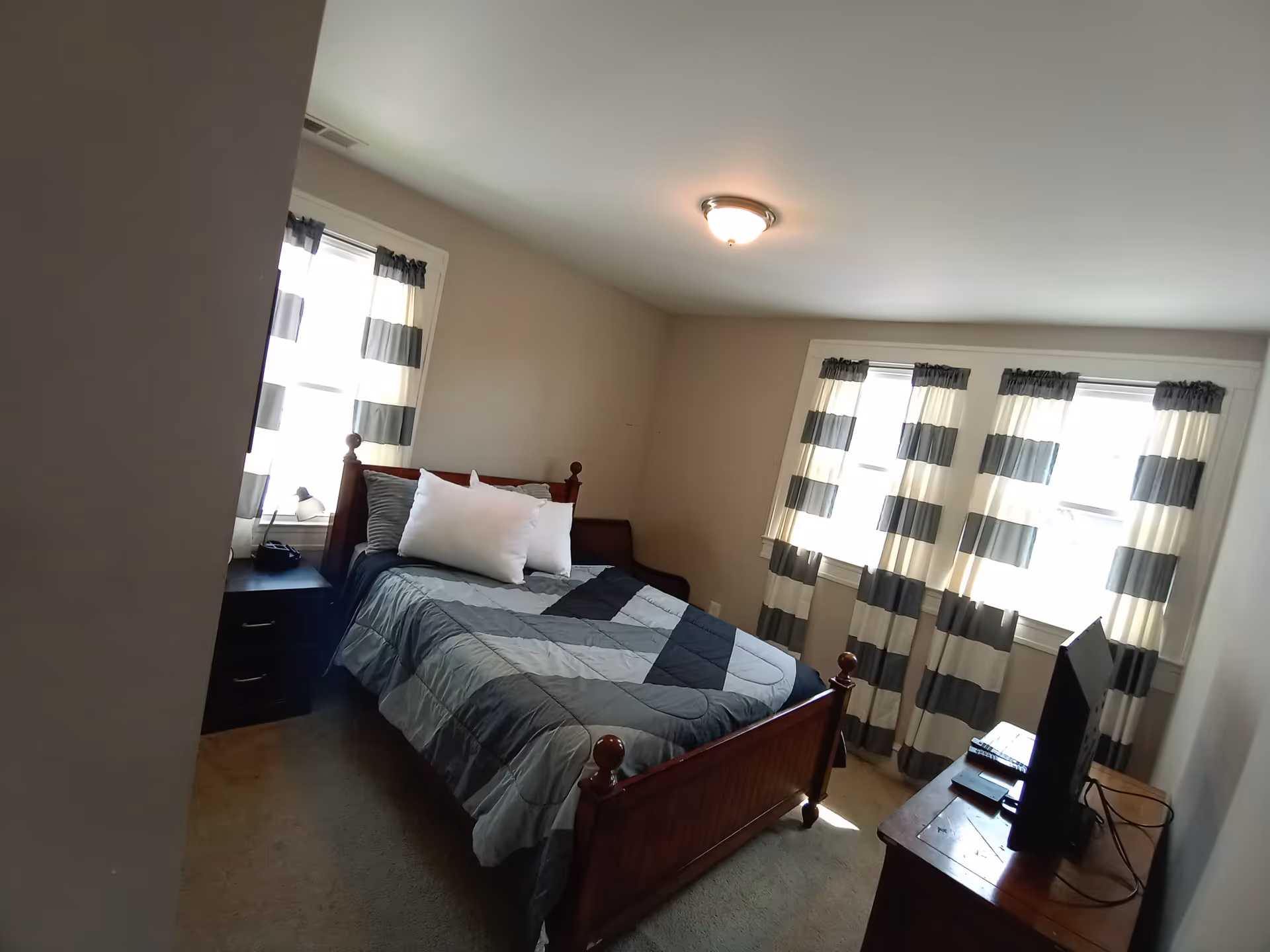 A furnished bedroom in an assisted living facility with a wooden bed, dresser, and striped curtains.