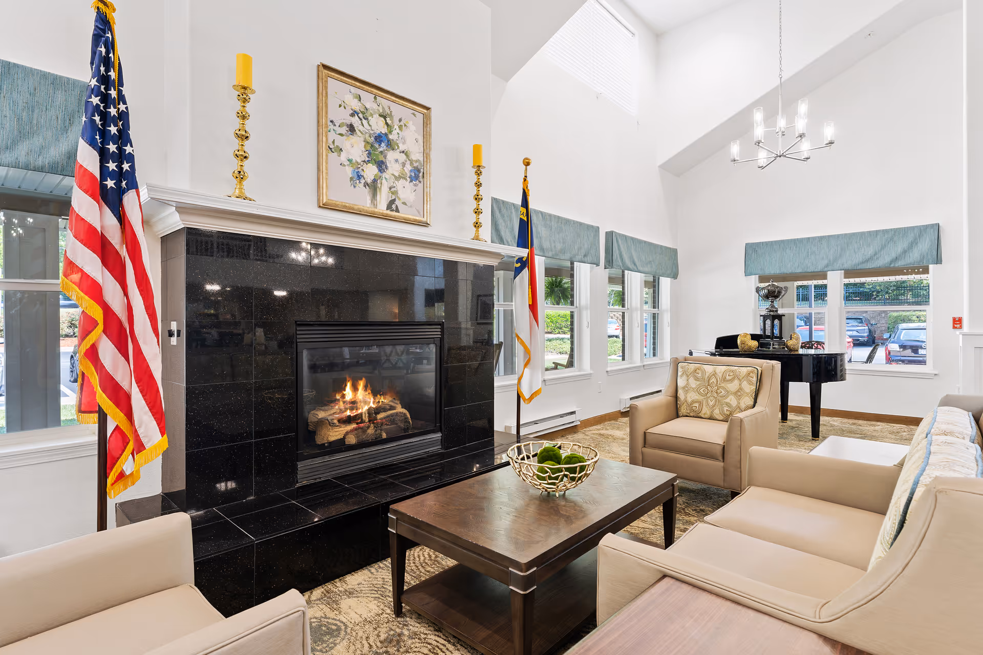 A bright and spacious living room with high ceilings featuring a lit black fireplace with a floral painting and two yellow candles on the mantel. The room includes beige armchairs and a sofa around a dark wooden coffee table with a decorative bowl. Two flags stand on either side of the fireplace. Large windows with blue valances let in natural light, and a black grand piano is visible near the windows.