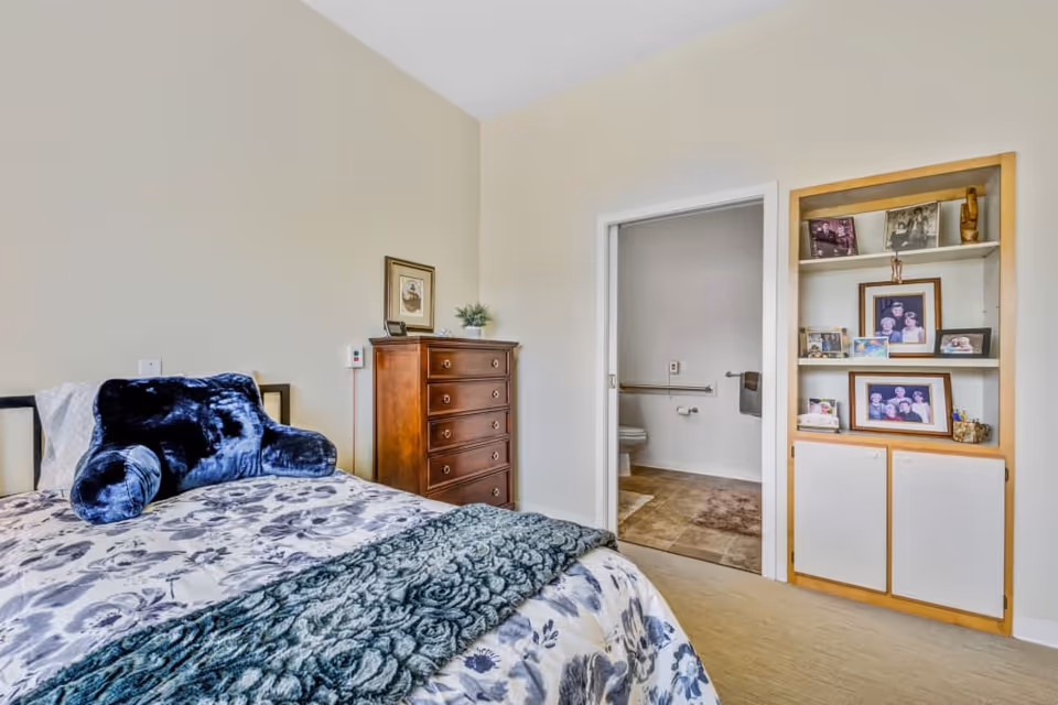 A bedroom in a senior living facility featuring a bed with floral bedding and a dark blue plush pillow, a wooden chest of drawers with a framed picture and a small plant on top, and an open doorway leading to a bathroom with a toilet and grab bars. To the right of the doorway is a built-in shelving unit displaying several framed family photos and small decorative items.
