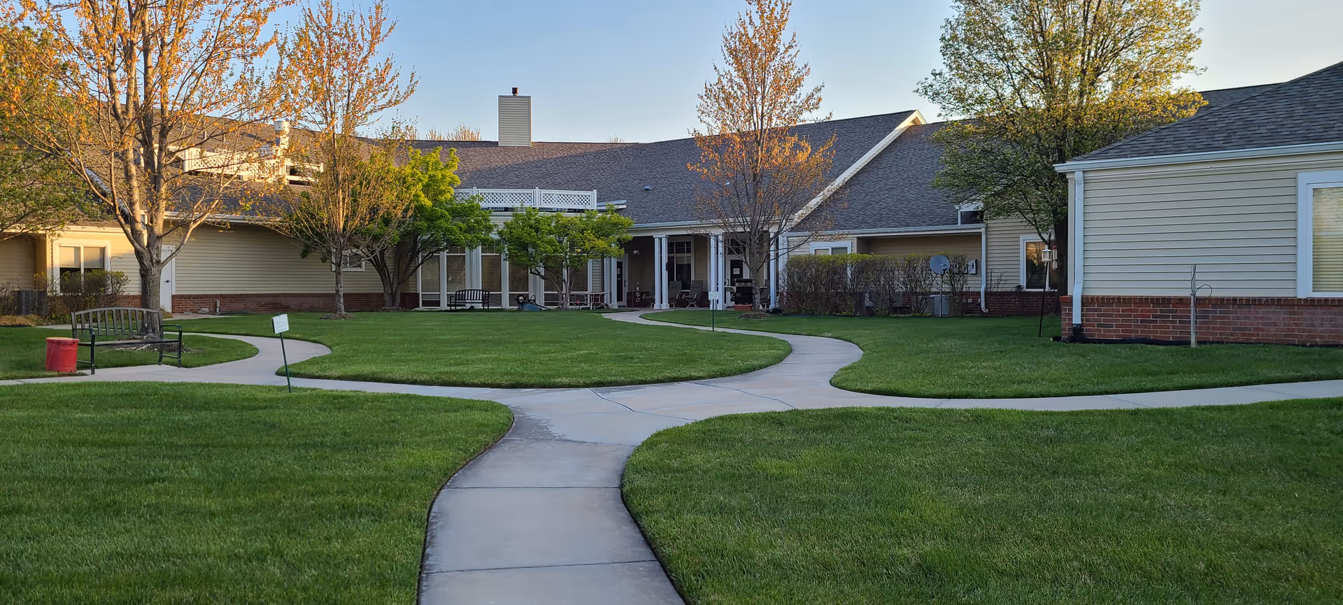 Courtyard with intersecting concrete walkways, green lawn, benches and a single-story residential building with trees.