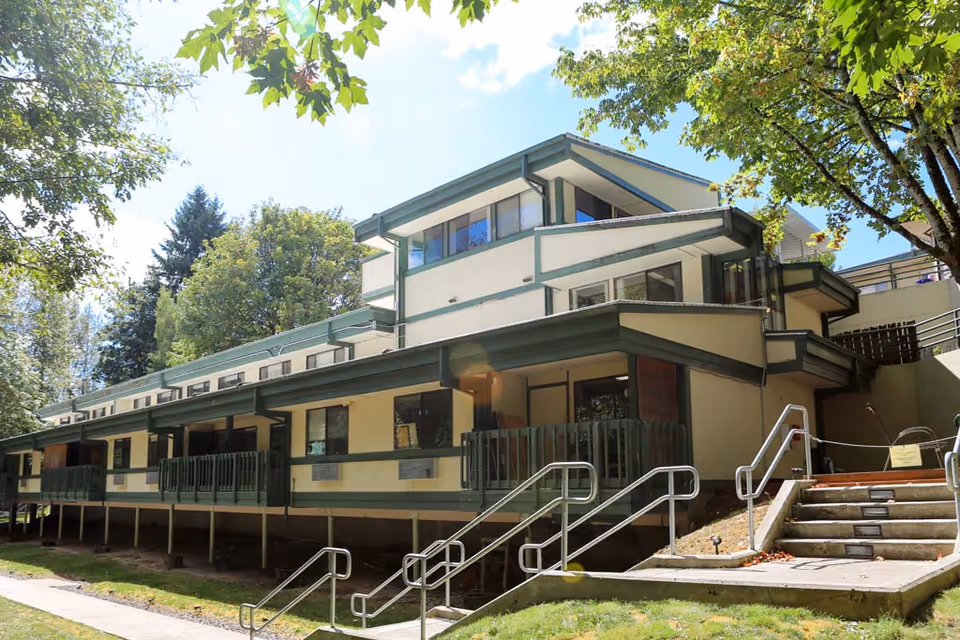 Exterior view of a multi-level building with green railings and trim, surrounded by trees and greenery under a blue sky with some clouds. There are stairs with metal handrails leading up to the entrance area.