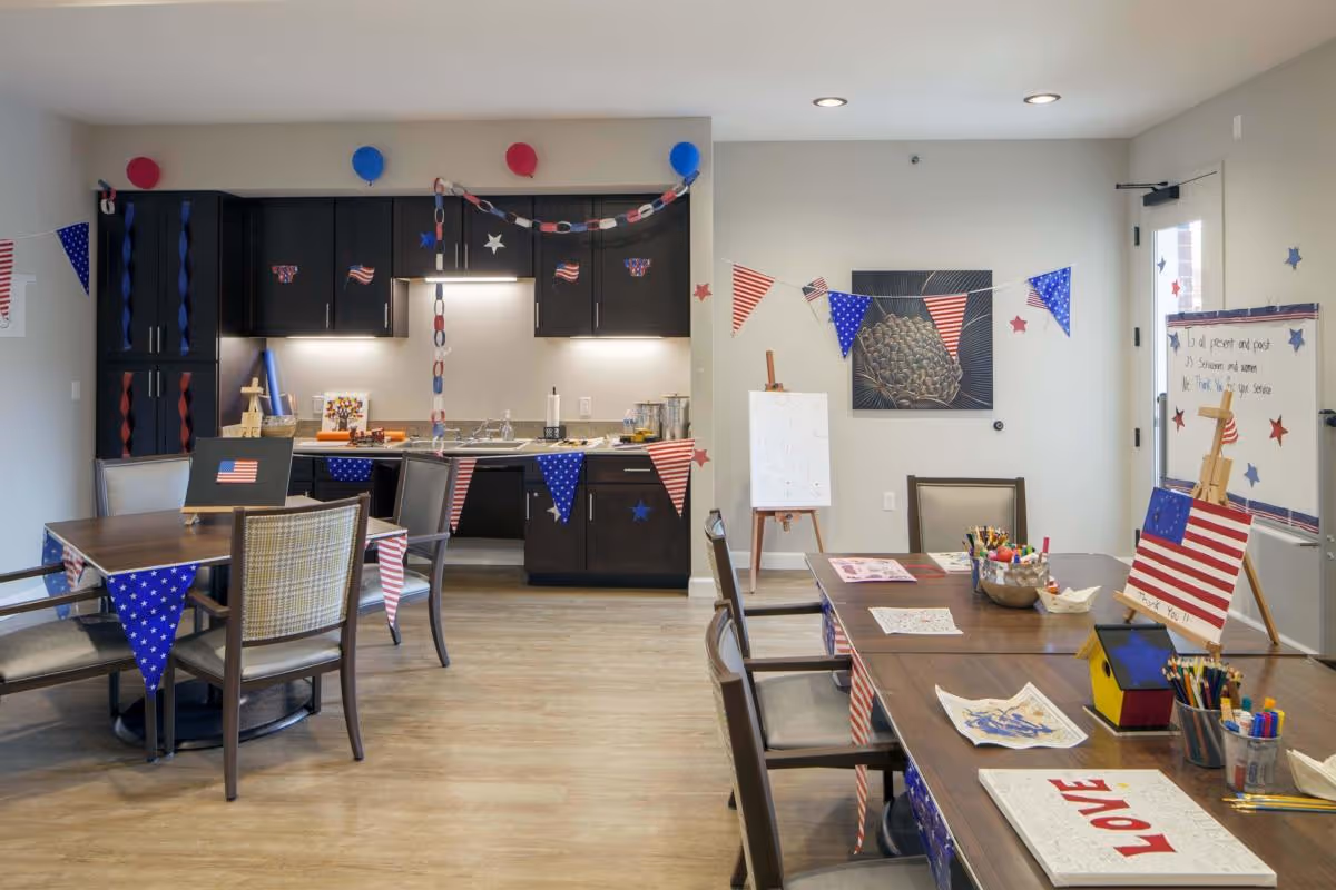 A decorated activity room with tables and chairs, featuring patriotic red, white, and blue bunting, paper chains, and small American flags. The room includes a kitchenette with dark cabinets and a countertop, and tables set up with art supplies, coloring sheets, and craft materials. The walls have festive decorations and a piece of artwork, and there is a whiteboard with writing on it near the door.