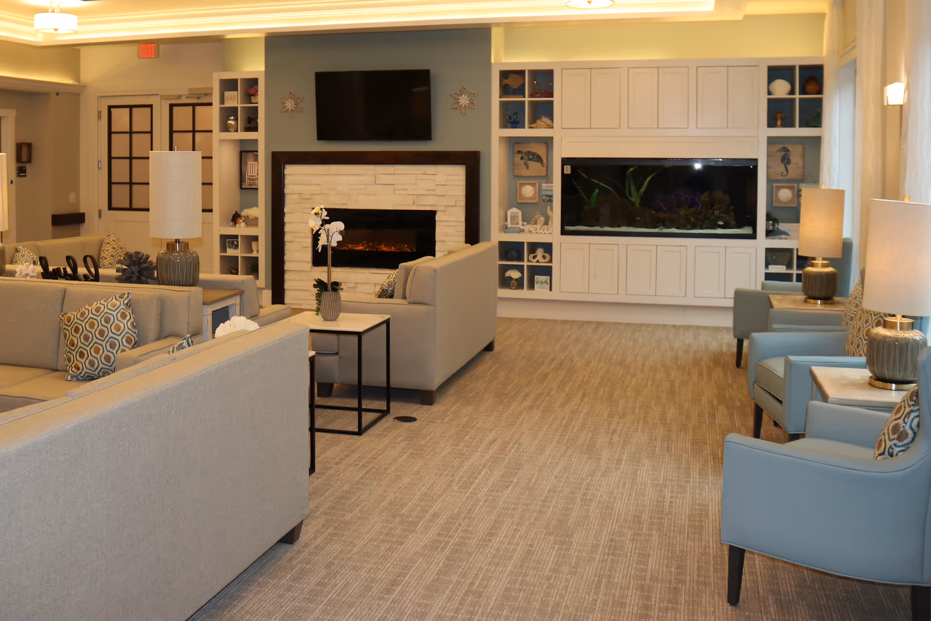 A cozy living room area in a senior living facility featuring beige sofas with patterned pillows, blue armchairs with matching pillows, side tables with lamps, a modern electric fireplace with a TV mounted above it, and a large built-in aquarium surrounded by decorative shelves with coastal-themed decor.
