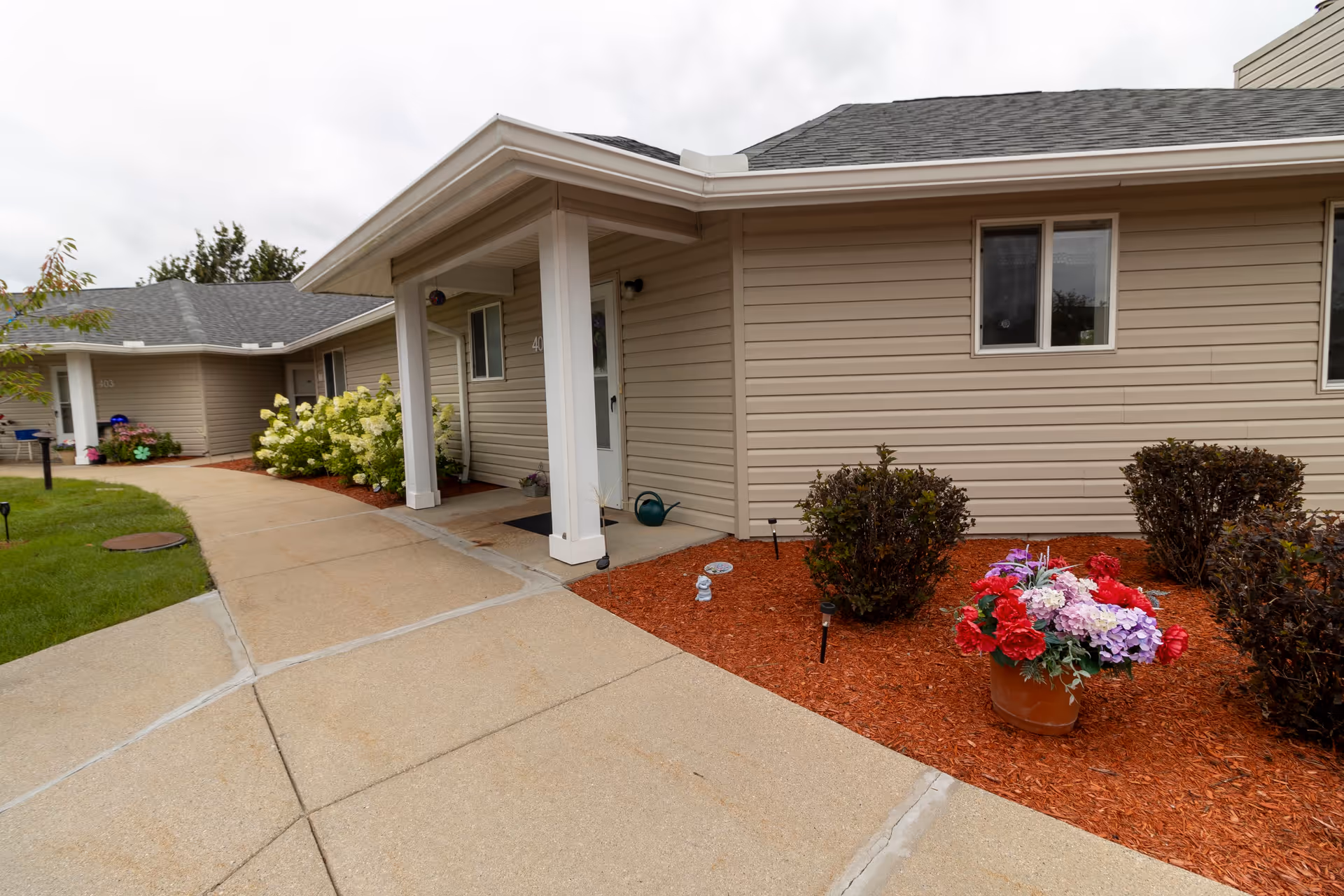 Exterior view of a beige single-story building with a covered entrance supported by white columns. There is a concrete walkway leading to the entrance, surrounded by landscaped areas with mulch, bushes, and a pot of colorful flowers. The sky is overcast.