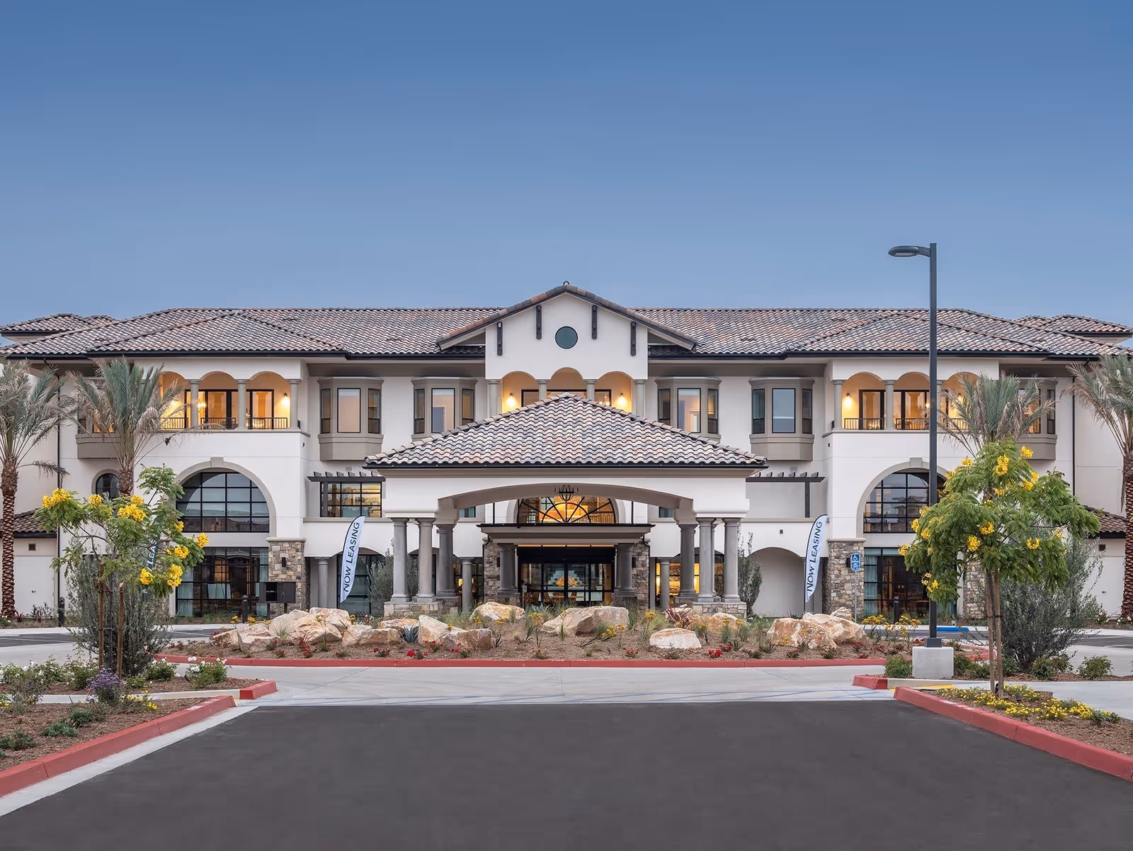 Front exterior view of Silvergate Rancho Bernardo facility with a tiled roof, arched windows, palm trees, and a landscaped entrance with rocks and flowers under a clear sky.