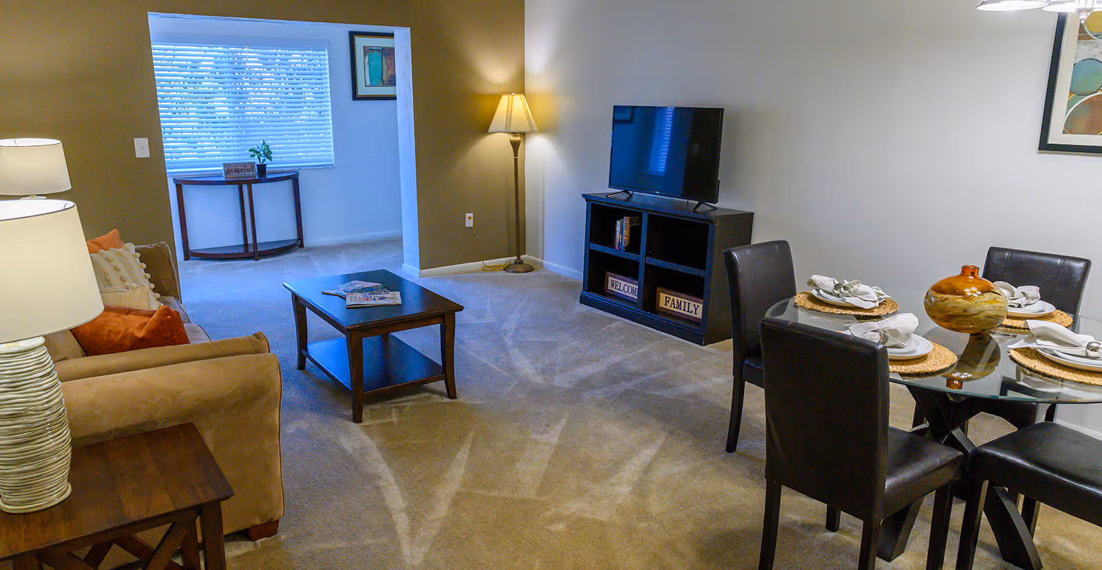 A cozy living room and dining area in a senior living facility. The living room features a beige sofa with orange and white pillows, a wooden coffee table, and a TV on a black stand with decorative signs. The dining area has a round glass table set with four black chairs and place settings. There are two lamps providing warm lighting, and a window with blinds in the background.