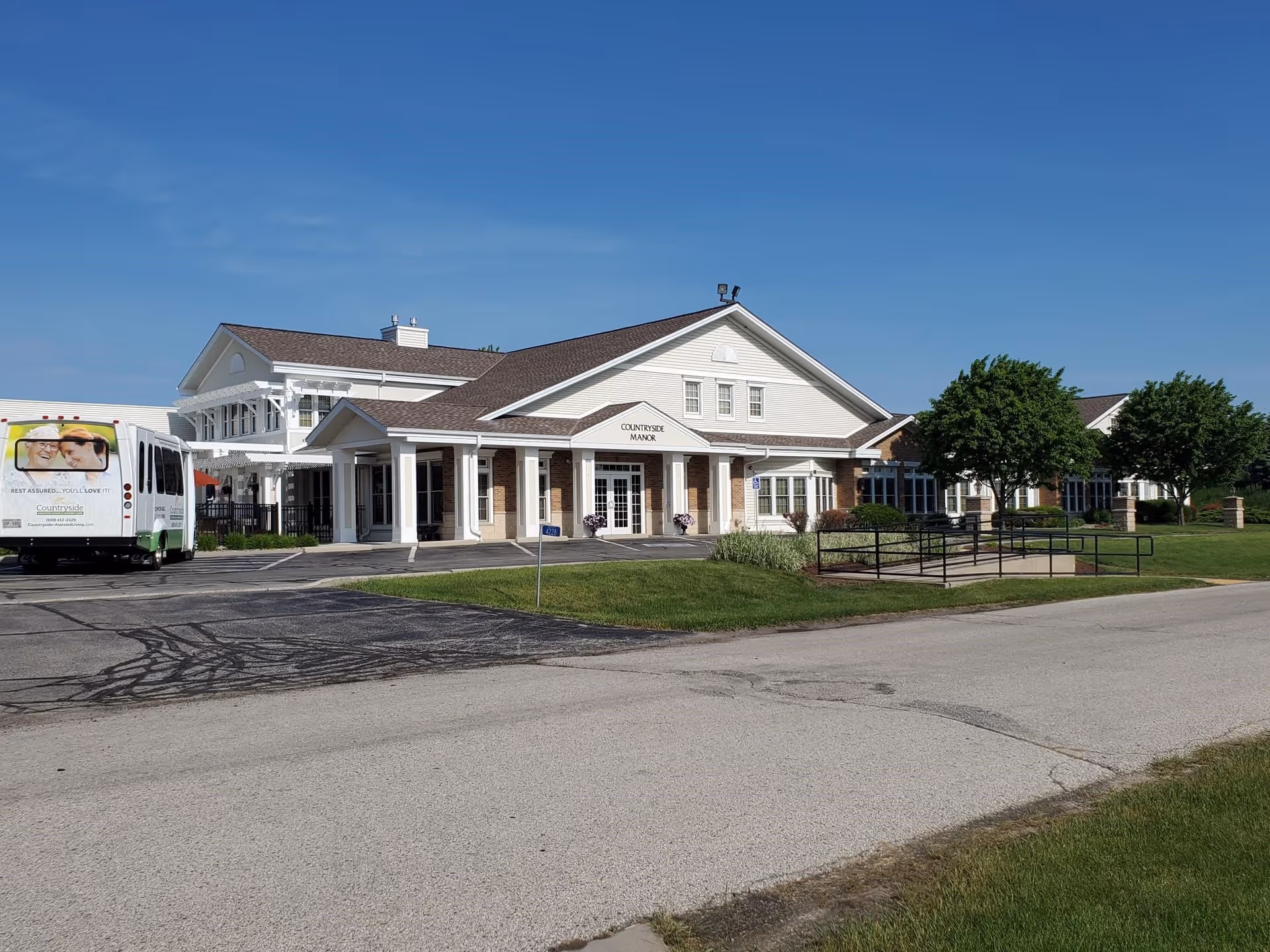 Exterior view of Countryside Manor, a large white building with a brown roof, featuring a covered entrance with columns, surrounded by green lawns and trees under a clear blue sky. A shuttle bus with images of smiling elderly people is parked on the left side.