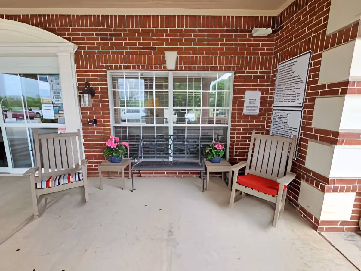 Covered outdoor seating area with two wooden chairs, one with a red cushion, a metal bench, two small tables with pink flowers in pots, and a brick wall with windows and posted notices.