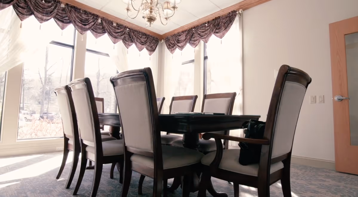 A bright dining room with a dark wooden dining table surrounded by eight upholstered chairs. Large windows with sheer white curtains and decorative valances allow natural light to fill the room. A chandelier hangs from the ceiling, and a wooden door with a frosted glass panel is visible on the right.