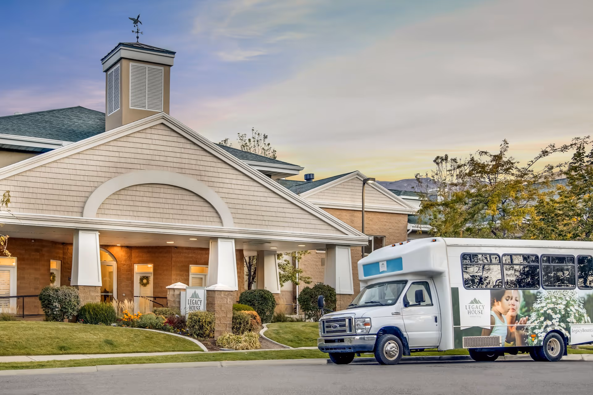 Front entrance of the Legacy House of Bountiful senior living facility with a branded shuttle bus parked in front.