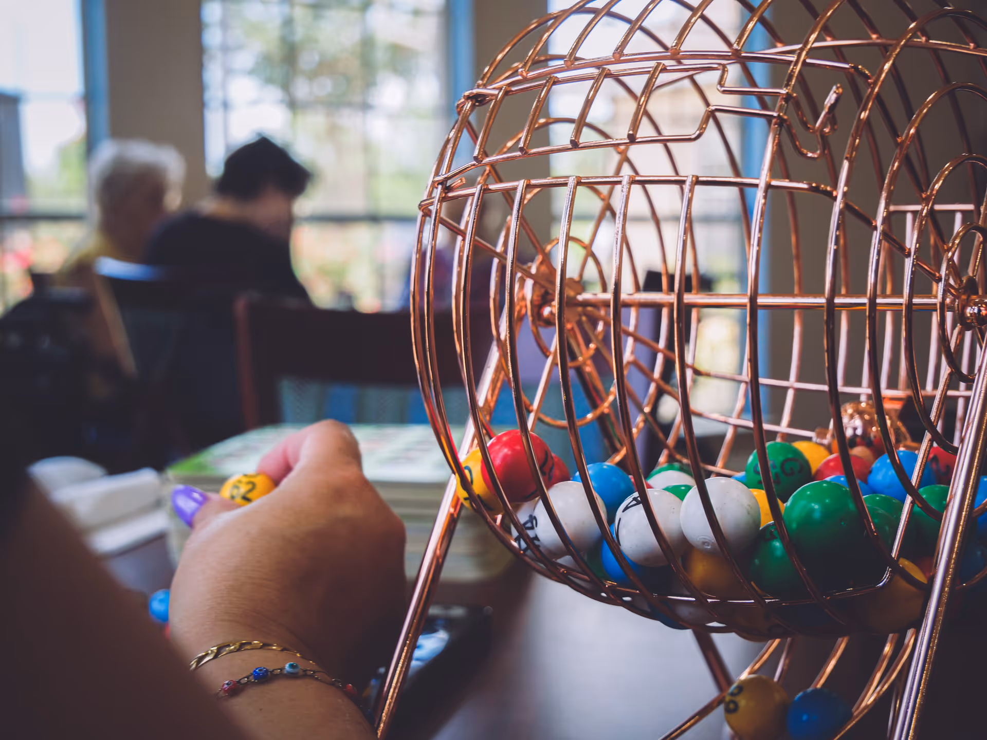 A bingo cage filled with colorful balls on a table as a person marks a card in a communal room.