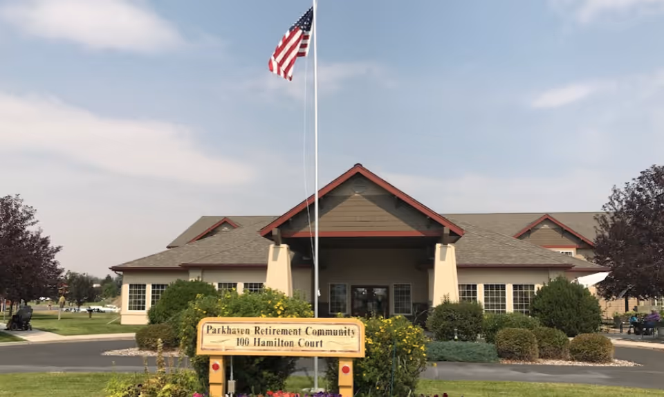 Front exterior view of Parkhaven Retirement Community building with a peaked roof, surrounded by bushes and trees, an American flag on a flagpole, and a sign in front displaying the community name and address.