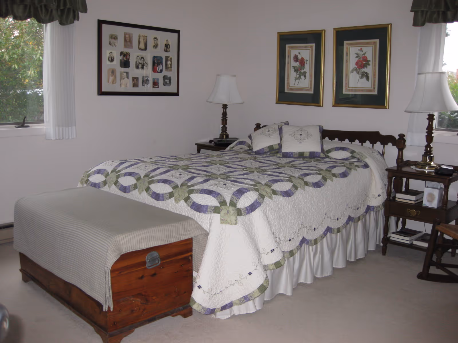 A bedroom with a bed covered in a white quilt with green and purple circular patterns, two matching pillows, and a wooden headboard. There are two wooden nightstands on either side of the bed, each with a lamp. A wooden chest with a grayish cover is at the foot of the bed. The room has light-colored walls with framed pictures hanging above the bed and a window with white curtains on each side.
