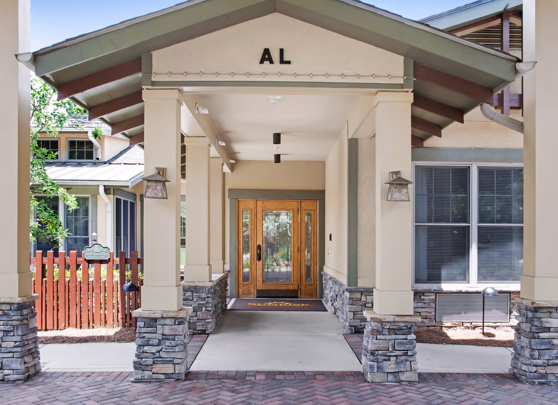 Covered entrance to a building with stone pillars and a wooden door with glass panels. The letters 'AL' are displayed above the entrance. There is a red wooden fence and windows with blinds on the right side.