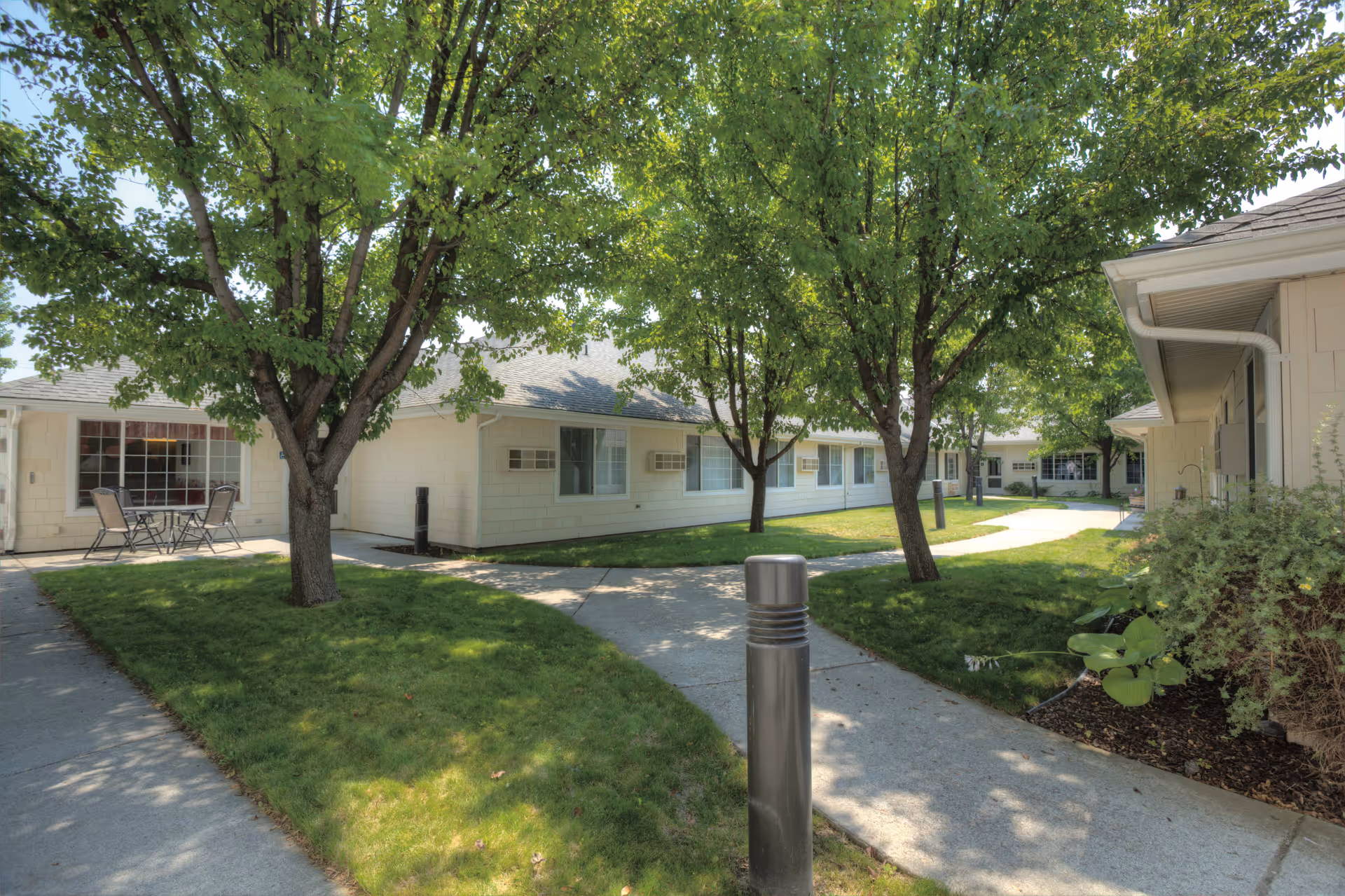 Outdoor courtyard area at Eagle Springs Memory Care with paved walkways, green grass, several trees providing shade, and a small patio area with a table and chairs next to a single-story building.