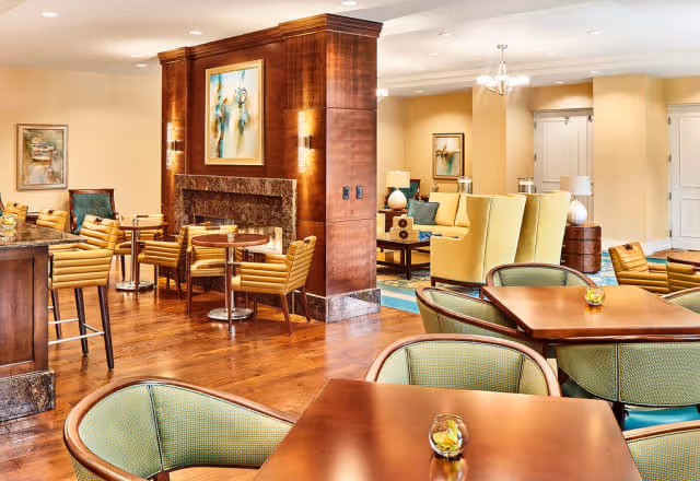 A spacious and warmly lit common area with wooden floors, multiple seating arrangements including tables with green cushioned chairs and yellow armchairs, a central wooden column with a fireplace, and framed artwork on the walls.