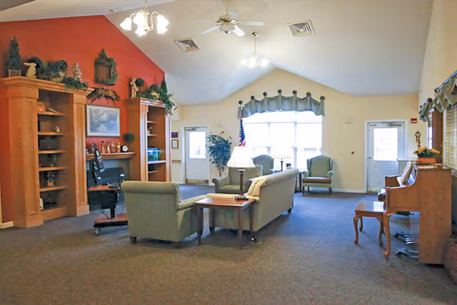 A spacious living room area in a senior living facility with beige walls and carpeted floor. The room features a green sofa and armchair arranged around a wooden coffee table with a lamp. There is a wooden entertainment center with decorative items and a piano with a bench on the right side. Large windows with a valance allow natural light to fill the room. Ceiling fans and light fixtures hang from the vaulted ceiling.