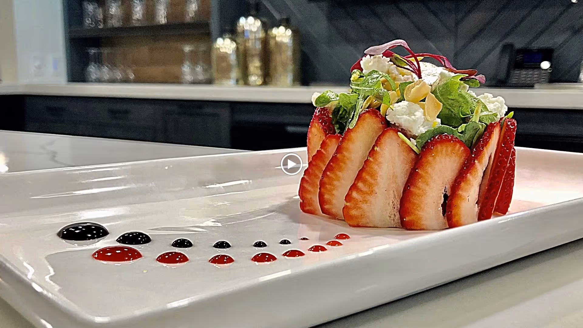 A gourmet salad dish presented on a white rectangular plate, featuring a ring of sliced strawberries surrounding mixed greens, goat cheese, and almond slices, with decorative drops of black and red sauce on the plate. The background shows a modern kitchen counter with jars and containers.