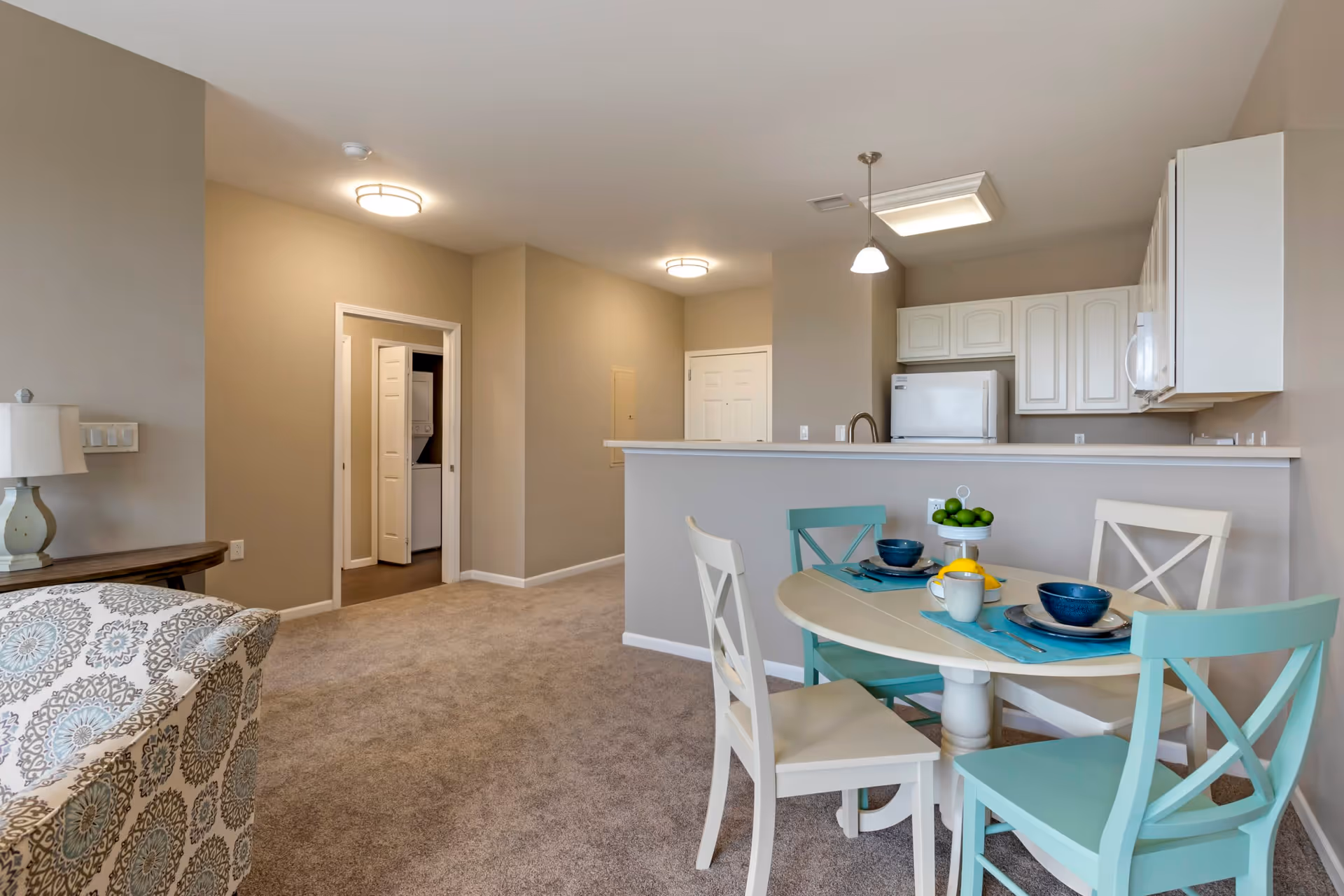 Interior view of a senior living apartment showing a small dining area with a round table set for two with blue and white chairs, a kitchen with white cabinets and appliances in the background, beige walls, carpeted floor, and a glimpse of a living area with a patterned armchair and a lamp.