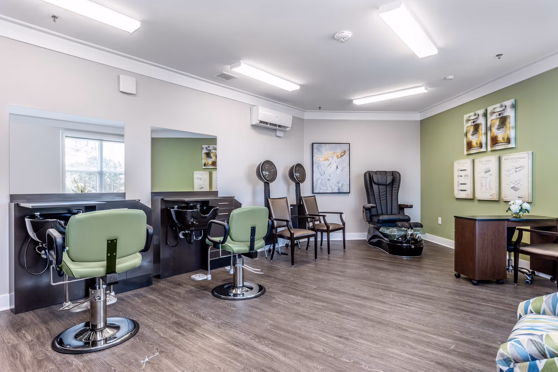 Interior view of a salon area in a senior living facility with two green salon chairs in front of black washbasins and mirrors, two hair dryers on stands, two wooden chairs, a black pedicure chair with a foot bath, a small desk with a chair, and framed artwork on the walls.