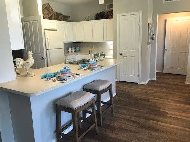 A modern kitchen area with a long white countertop island featuring two cushioned bar stools. The countertop is set with plates, bowls, glasses, and turquoise napkins. White cabinets line the walls, and there is a decorative rooster figurine on the counter. The floor is wooden, and there are two white doors visible in the background.