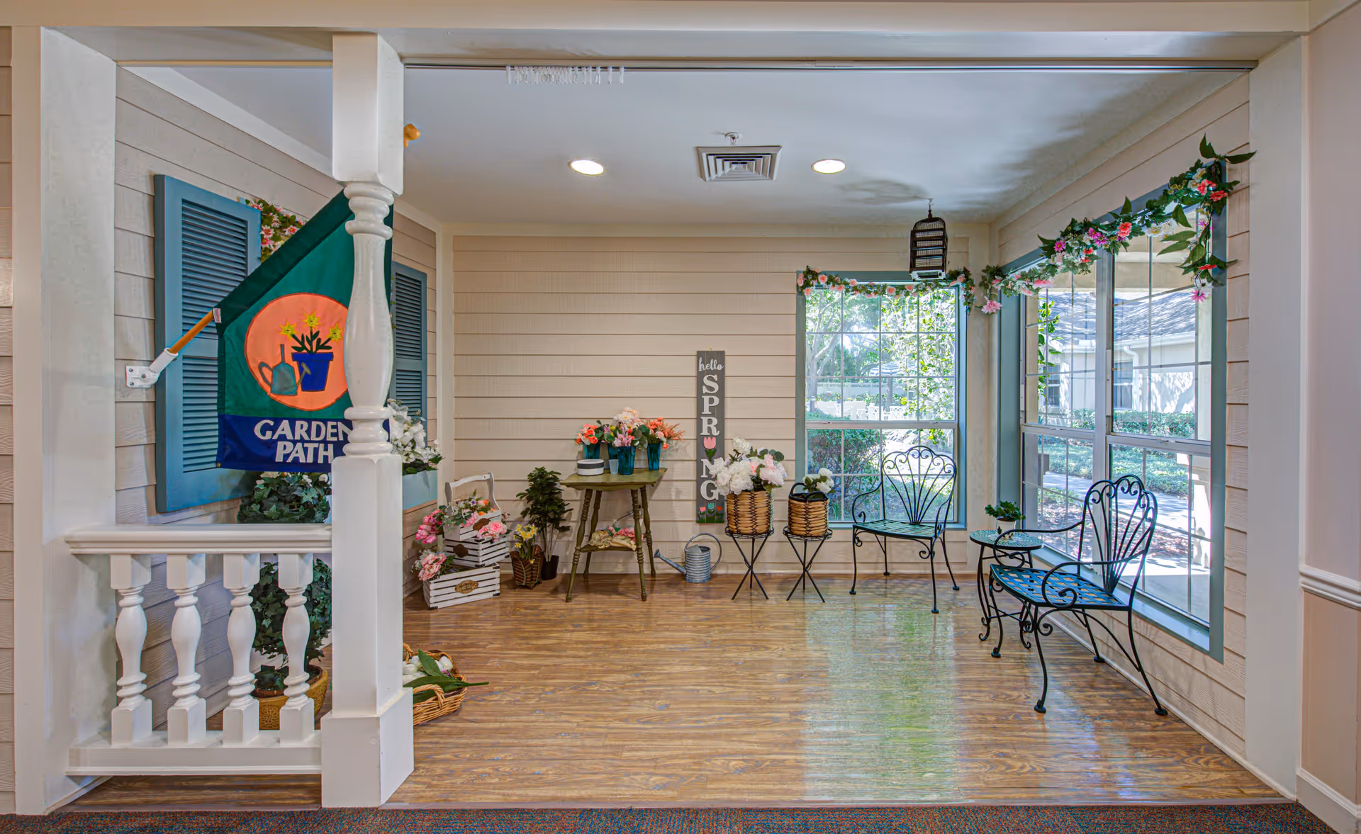 A bright indoor garden-themed sitting area with large windows letting in natural light. The space features a small green table with flower vases, two black wrought iron chairs, a matching bench, and various flower decorations including a hanging garland and potted plants. A sign on the wall reads 'hello SPRING'. A green flag with the words 'GARDEN PATH' is mounted on the left side near a white railing.