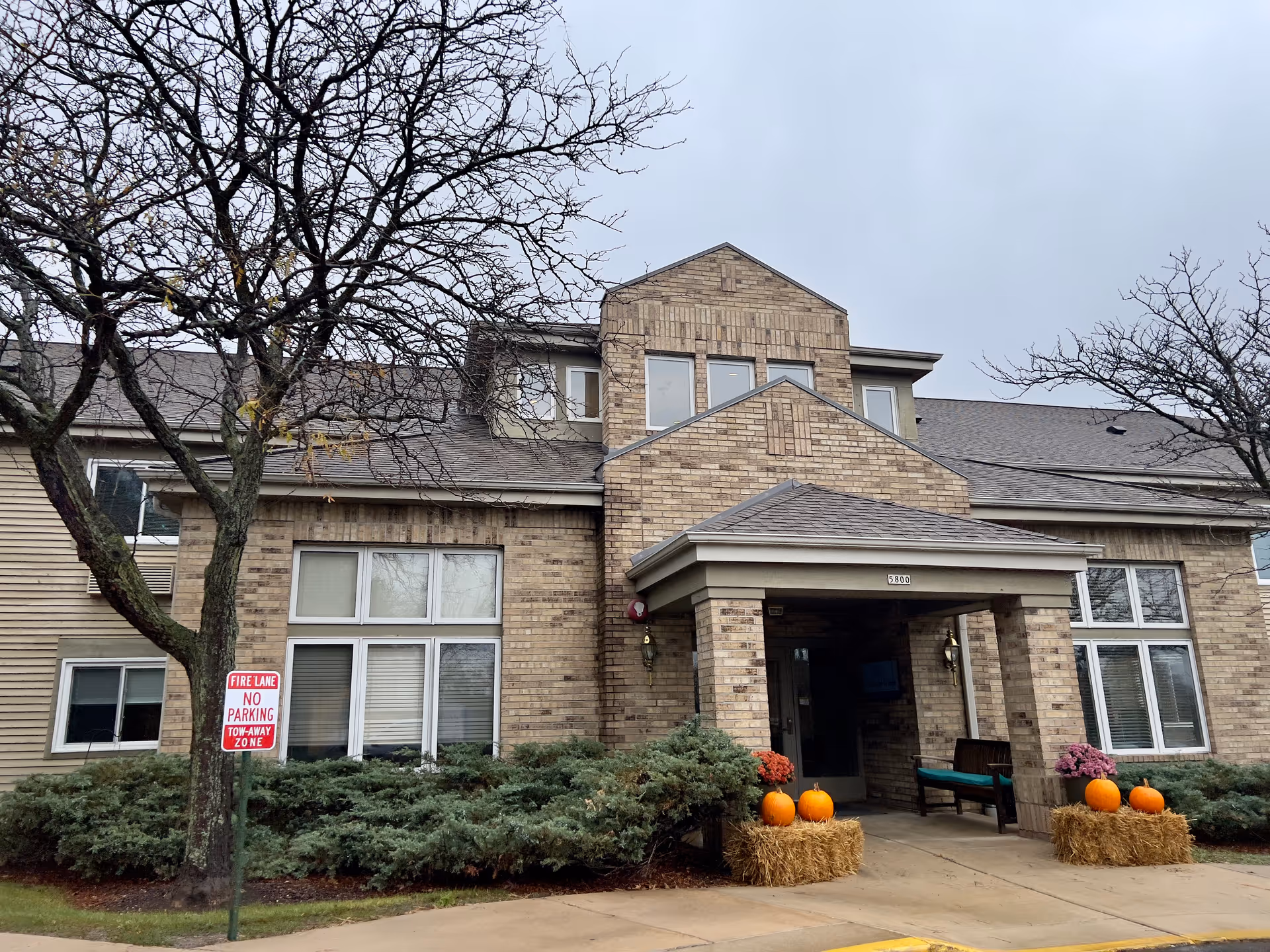 Exterior front view of a brick assisted living facility building with a covered entrance. There are two leafless trees and bushes in front, along with hay bales decorated with pumpkins and flowers near the entrance. A fire lane no parking sign is visible on the left side.
