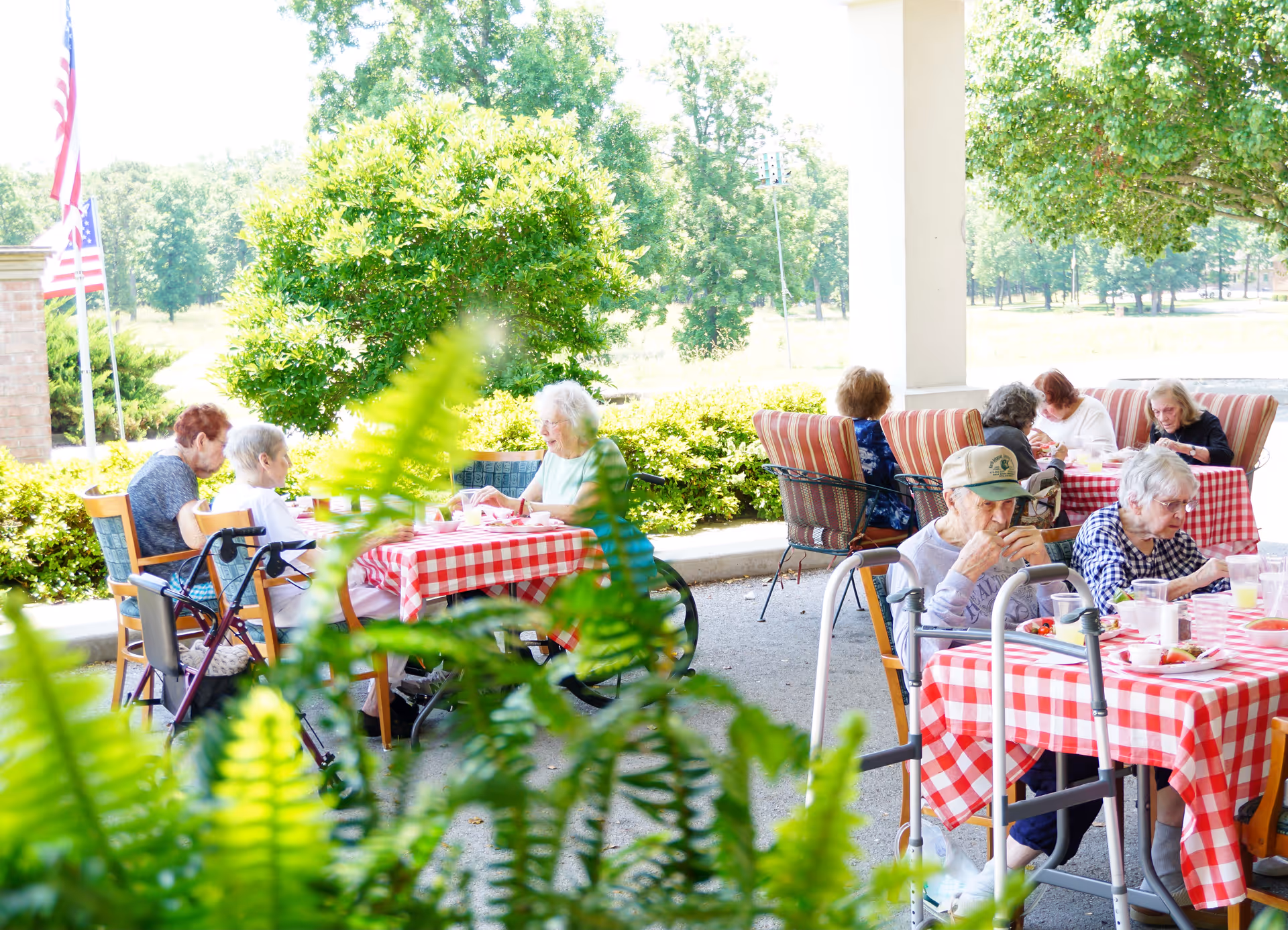 Several elderly people sitting at outdoor tables covered with red and white checkered tablecloths, enjoying a meal together on a patio surrounded by greenery and trees.