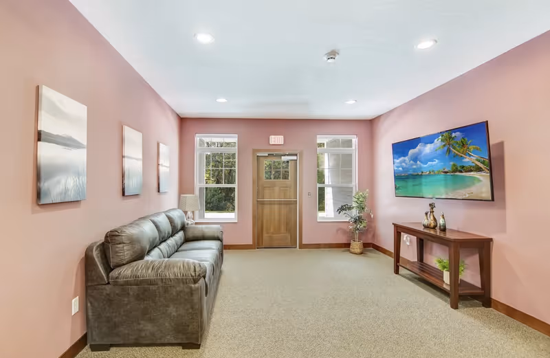 A cozy living room with pink walls, a gray leather sofa on the left, three landscape paintings above the sofa, two windows flanking a wooden door at the back, a flat-screen TV mounted on the right wall displaying a tropical beach scene, and a wooden console table beneath the TV with decorative items and a potted plant.