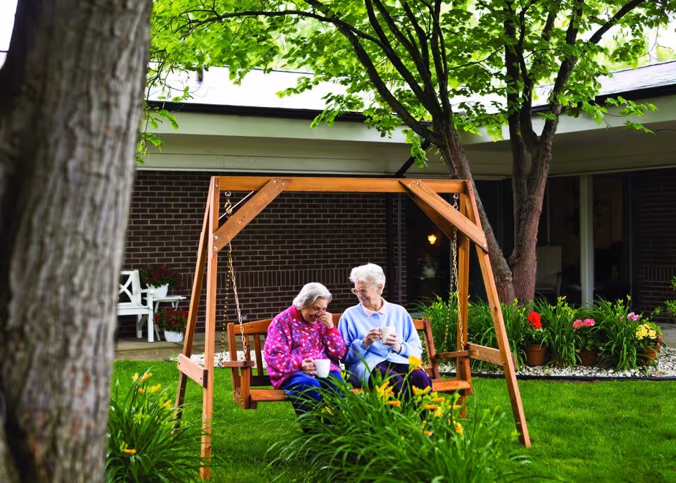 Two elderly women sitting on a wooden swing in a garden area outside a senior living facility, enjoying drinks and sharing a laugh. The background shows a brick building with windows and a variety of green plants and flowers around the swing.