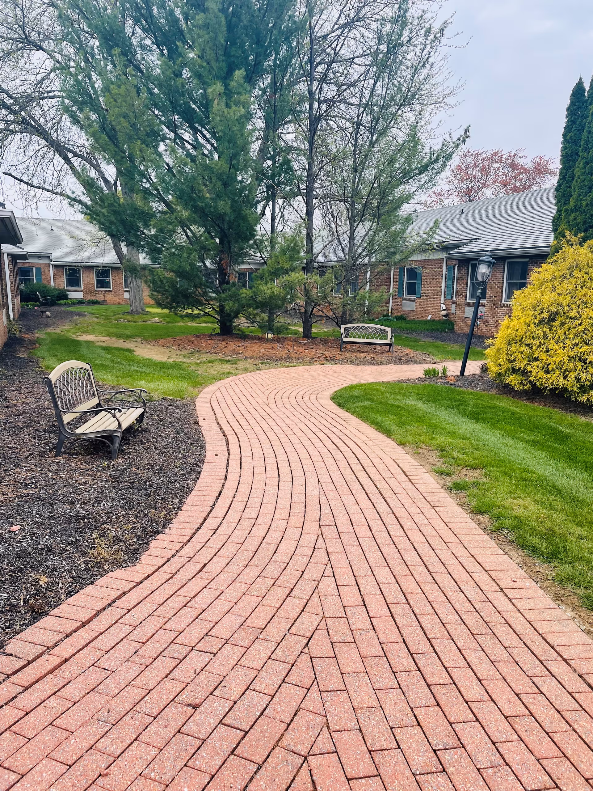Curved brick walkway in an outdoor garden area of a senior living facility, flanked by green grass, trees, bushes, and benches, with single-story brick buildings in the background under a cloudy sky.