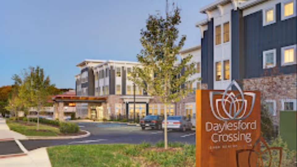 Exterior view of Daylesford Crossing senior living facility during early evening with a landscaped entrance, parking area, and a large sign displaying the facility's name and logo in the foreground.