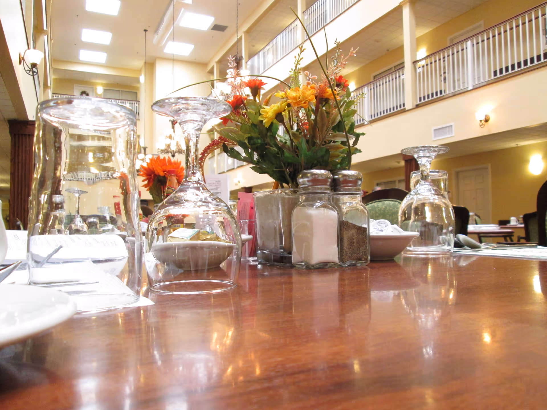 Close-up view of a dining table set with upside-down wine glasses, salt and pepper shakers, a small bowl, and a vase with colorful flowers in a spacious dining room with high ceilings and a balcony railing on the upper floor.