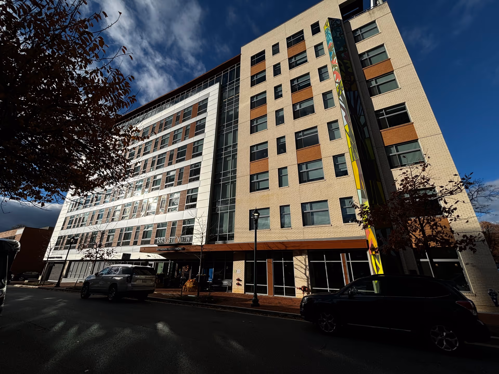 Exterior view of a multi-story modern building with large windows and a colorful vertical mural on one side. There are cars parked along the street in front of the building, and some trees with autumn leaves are visible. The sky is partly cloudy with patches of blue.
