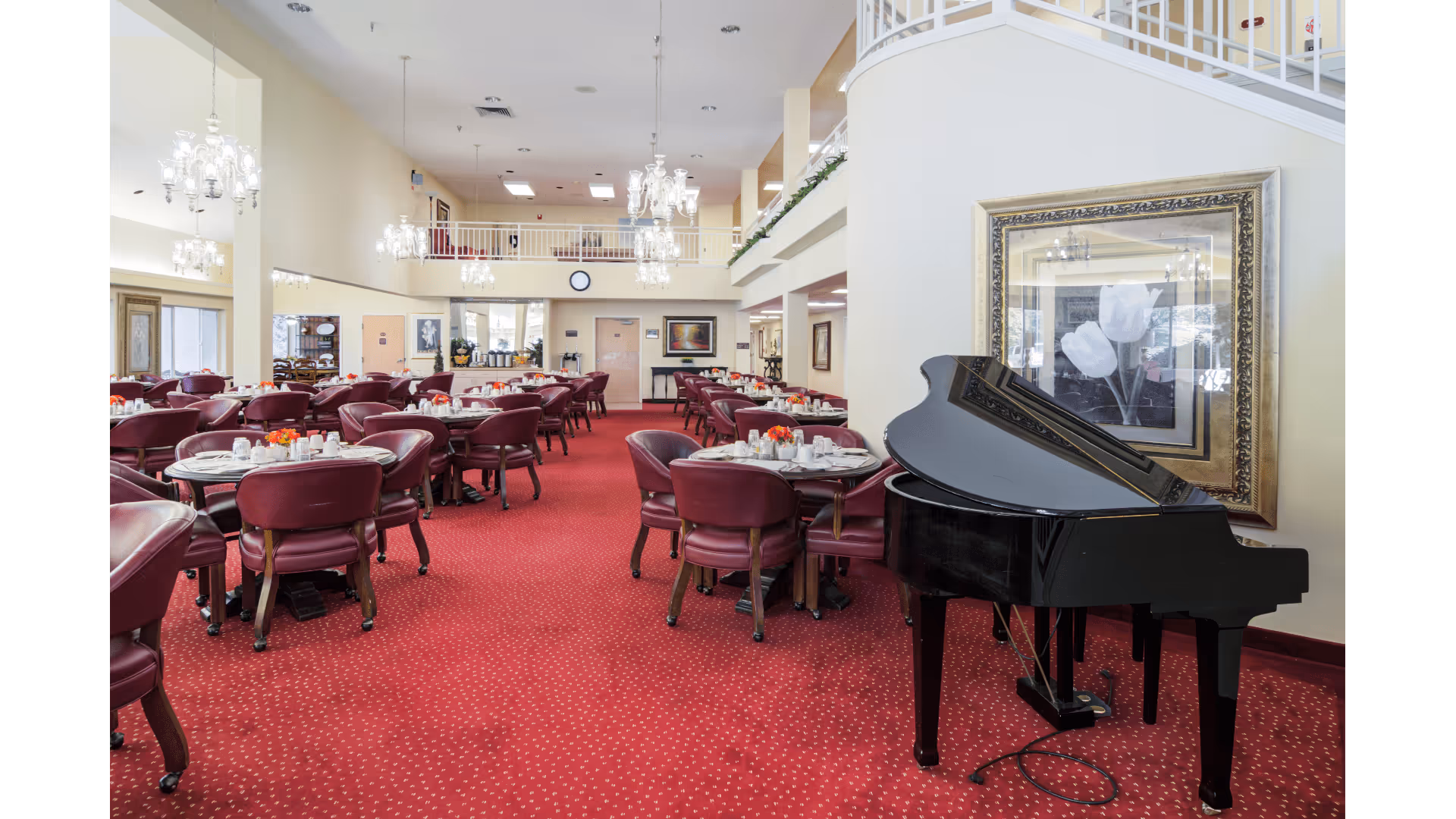 Spacious red-carpeted dining room with multiple round tables and burgundy chairs, chandeliers, and a black grand piano by a framed picture.