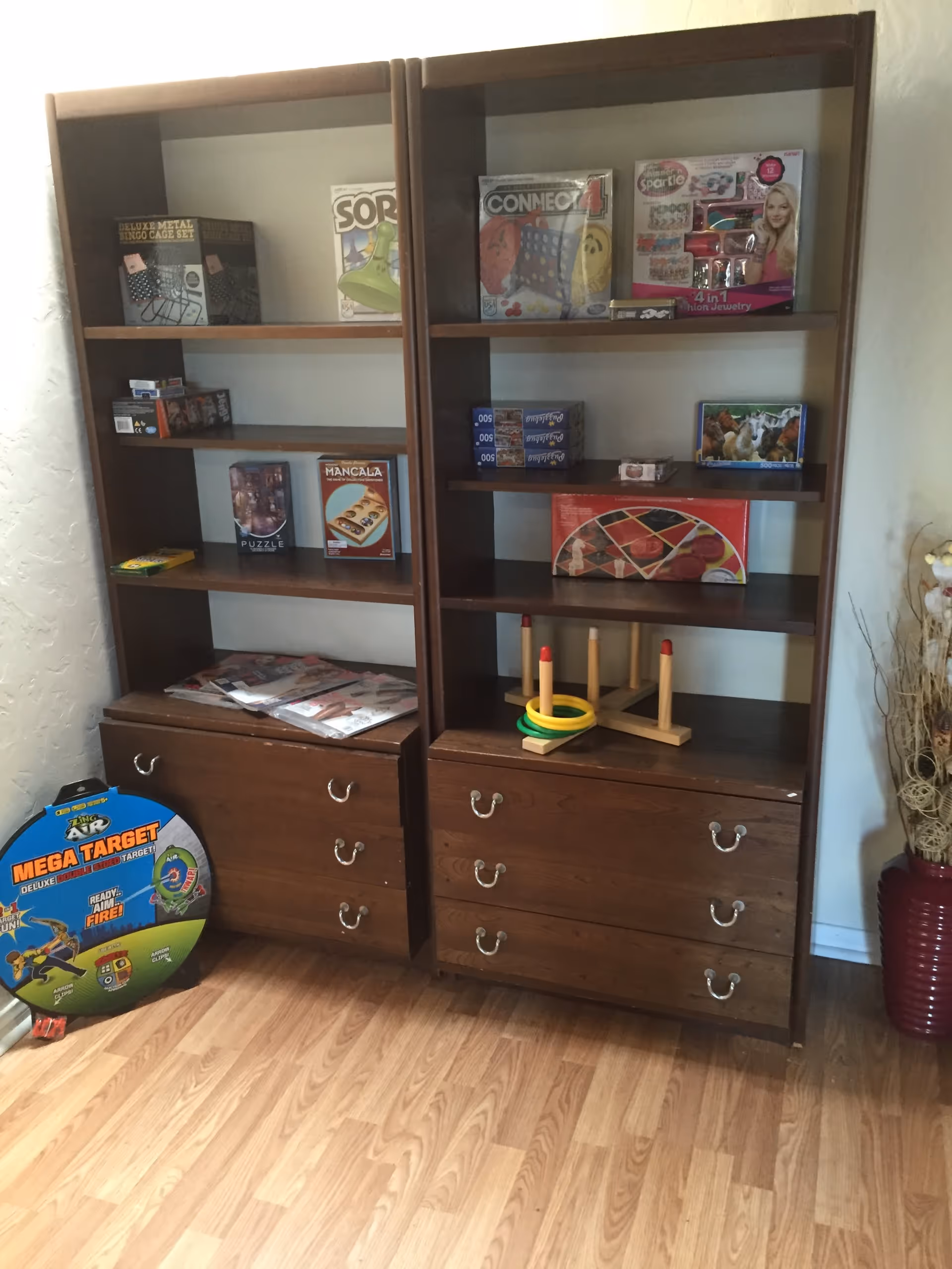 Two wooden shelving units with drawers at the bottom, containing various board games, puzzles, and a ring toss game. A large circular target game is leaning against the left shelving unit. The floor is wooden, and there is a decorative vase with dried plants on the right side.