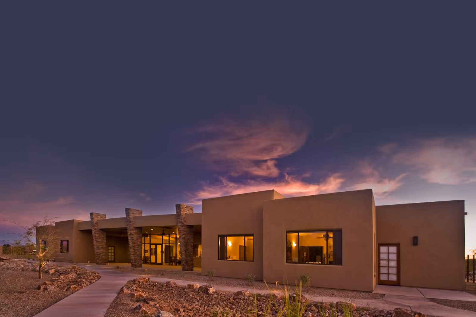 Exterior view of Via Elegante Assisted Living Sierra Vista Highlands building at dusk, featuring modern architecture with stone pillars, large windows with warm interior lighting, and a paved walkway surrounded by desert landscaping.