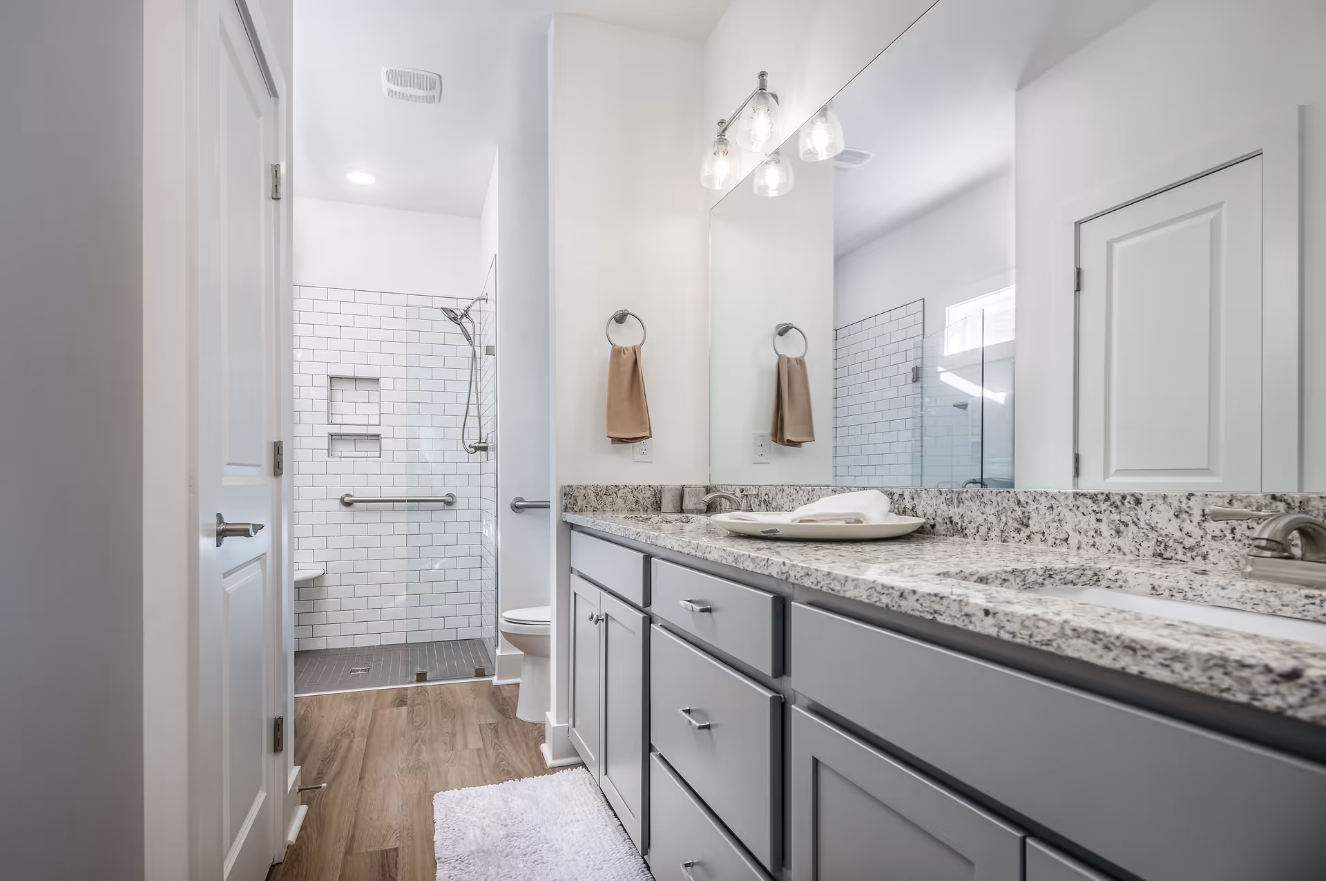 Modern bathroom with a granite countertop featuring two sinks, gray cabinets, a large mirror, and three glass light fixtures above. The floor is wood-style, and there is a white rug in front of the vanity. In the background, a walk-in shower with white subway tiles, a glass door, and a mounted showerhead is visible. A beige towel hangs on a ring next to the mirror.
