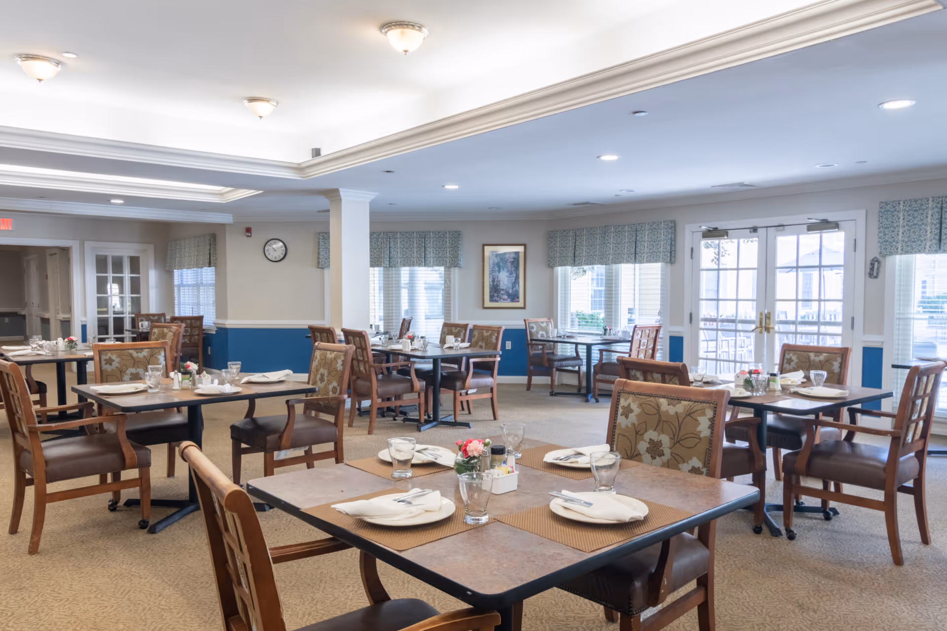 A bright and spacious dining room in a senior living facility with multiple tables set with plates, glasses, napkins, and silverware. The room features wooden chairs with floral upholstery, large windows with patterned valances, and double glass doors leading outside. The walls are painted beige with a blue wainscoting, and there is a clock and framed artwork on the walls.