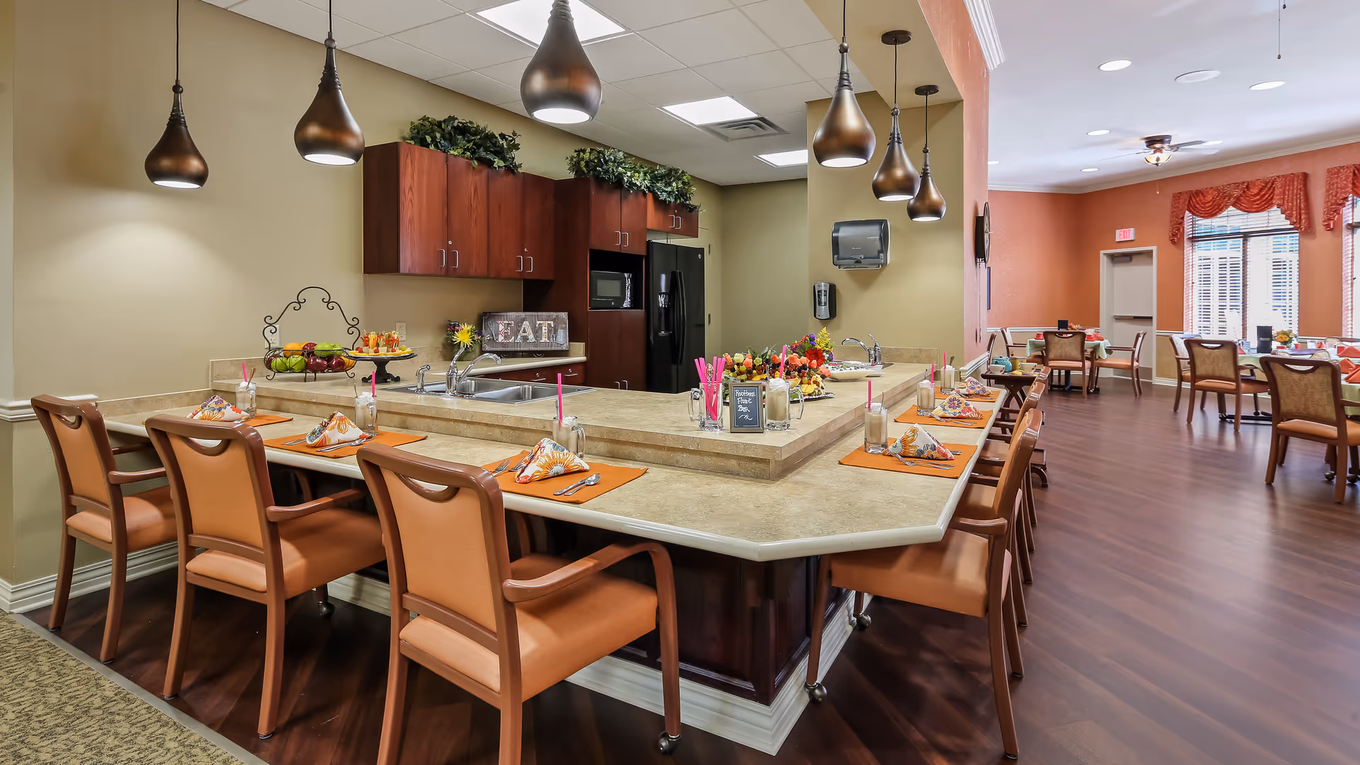Interior view of a dining area in a senior living facility featuring a large U-shaped counter with brown chairs and place settings. The counter has a sink, fruit displays, and decorative items. In the background, there are additional dining tables and chairs near windows with red valances, and warm lighting fixtures hang above the counter.