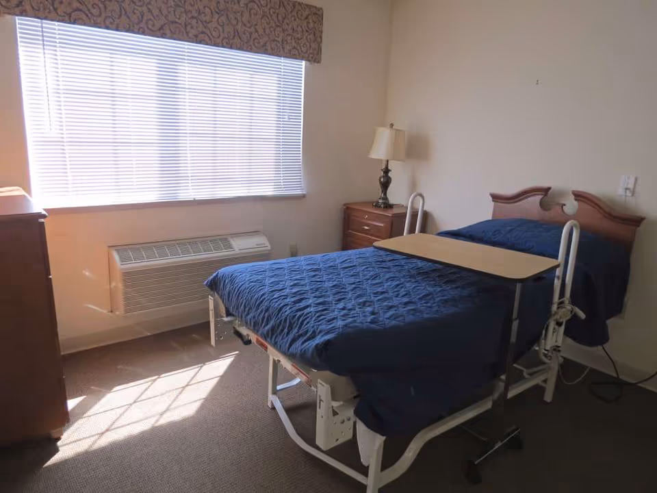 A small bedroom with a hospital-style bed covered in a dark blue quilt. The bed has side rails and an overbed table. There is a wooden nightstand with a lamp next to the bed, a window with blinds and a decorative valance, and a heating/cooling unit beneath the window. The room has beige walls and carpeted floor.