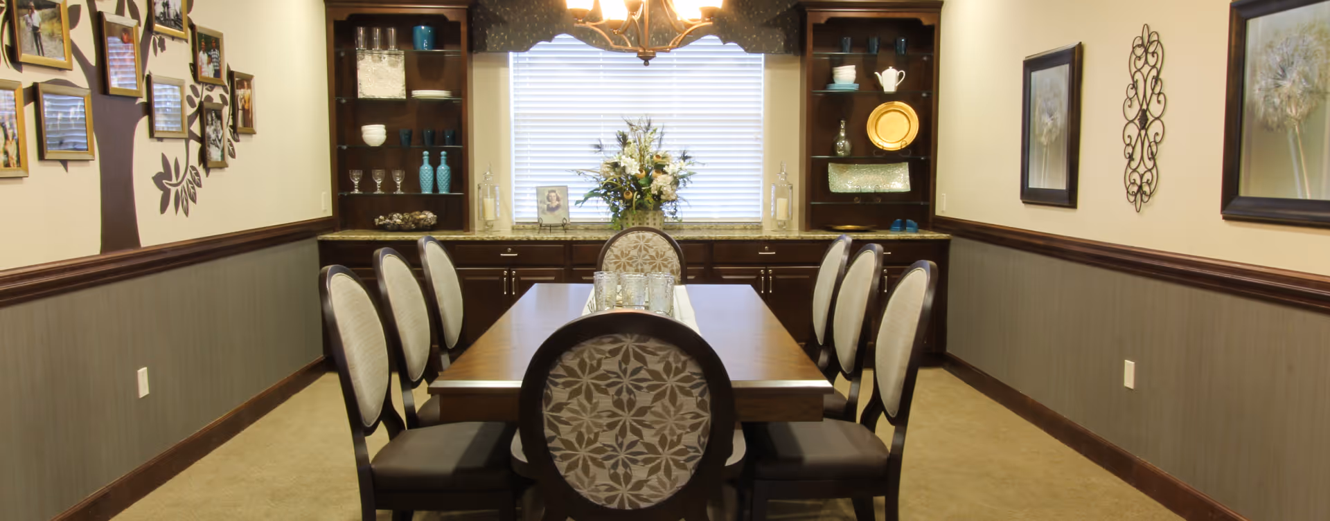 A formal dining room with a rectangular wooden table surrounded by eight upholstered chairs. The room features built-in dark wood cabinets with decorative dishes and glassware, a window with blinds, a chandelier overhead, and framed artwork on the walls.