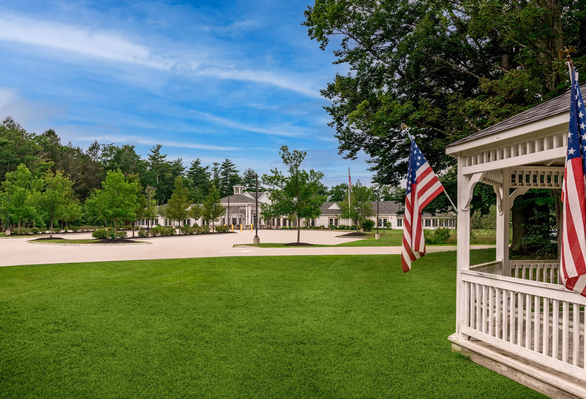View of a senior living facility with a large green lawn in the foreground, a white gazebo adorned with two American flags on the right, and a building with a cupola and multiple windows in the background surrounded by trees under a blue sky with some clouds.