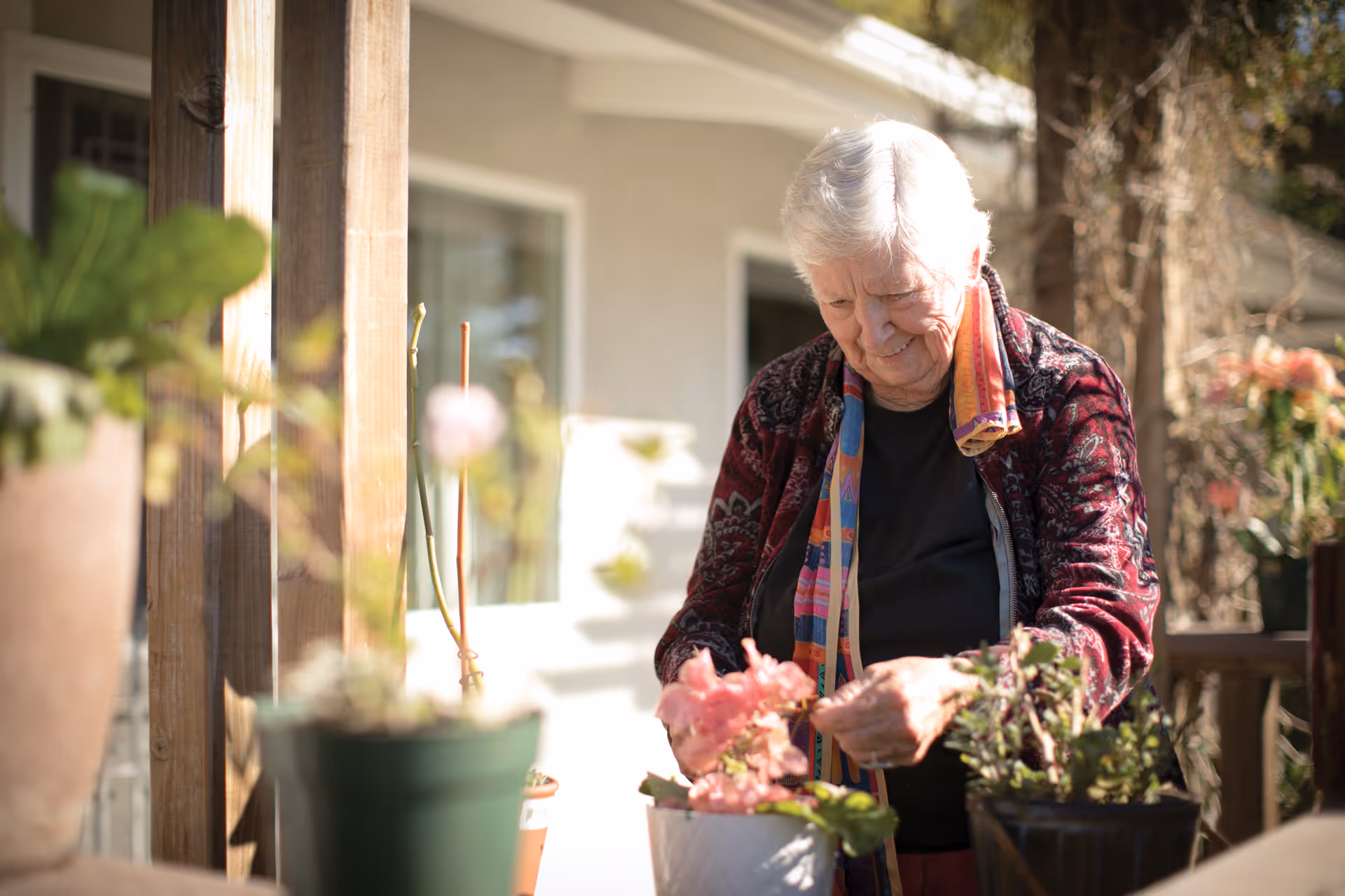 An elderly woman tending to potted plants on a wooden porch or patio area outside a building, with sunlight illuminating the scene and greenery around her.