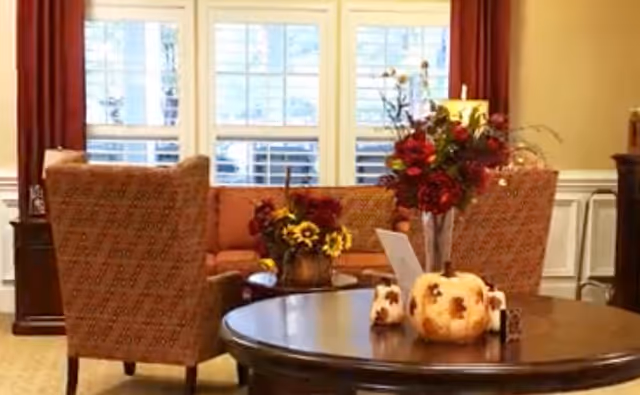 A cozy senior living room with patterned armchairs, a sofa, and a round wooden table decorated with autumn-themed pumpkins and a vase of flowers. Large windows with white shutters and red curtains allow natural light to fill the room.