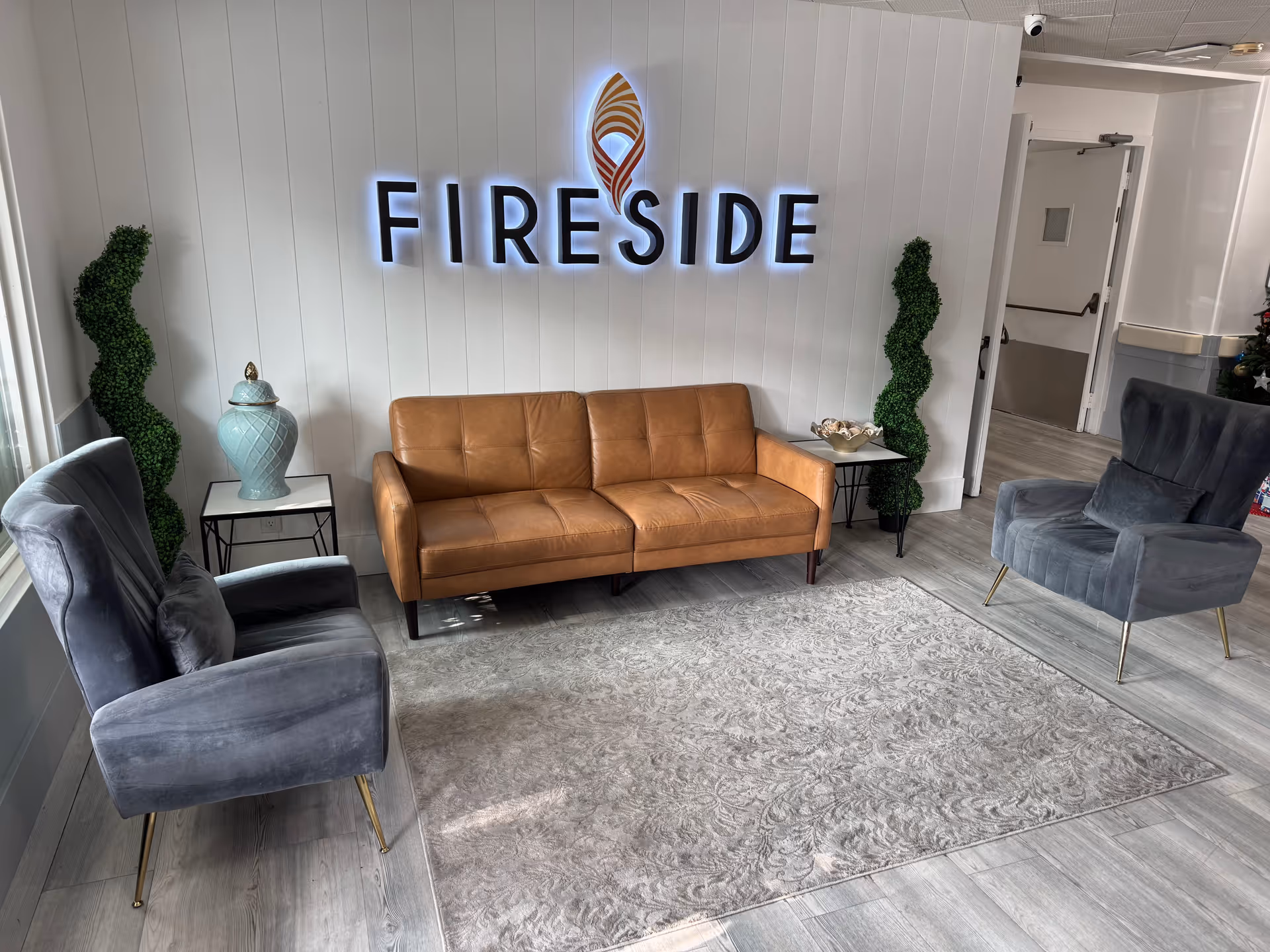 A modern lounge area with a brown leather sofa centered against a white paneled wall featuring the illuminated word 'FIRESIDE' and a flame logo above it. Two gray upholstered armchairs with gold legs are positioned on either side of the sofa. There are two small side tables with decorative items and two tall green topiary plants flanking the sofa. The floor is light wood with a large patterned area rug in front of the seating.