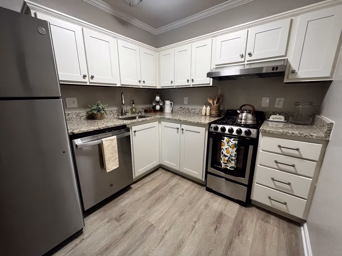 A modern kitchen with white cabinets, granite countertops, and stainless steel appliances including a refrigerator, dishwasher, and stove with a kettle on top. The kitchen has a light wood floor and various kitchen utensils and decor items on the counters.