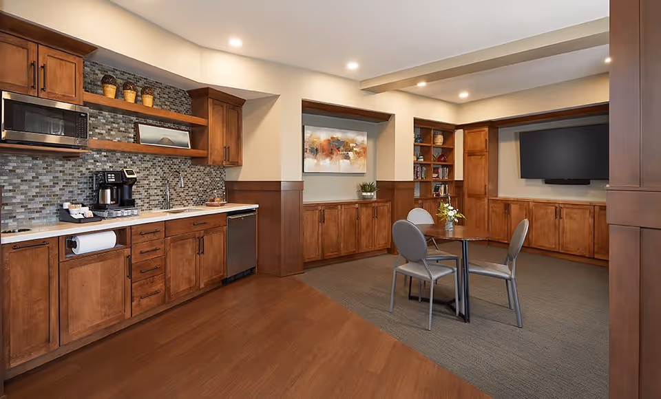 A cozy senior living common area featuring a kitchenette with wooden cabinets, a microwave, coffee maker, and a dishwasher. Adjacent to the kitchenette is a small round table with four chairs on a carpeted floor. The room has built-in wooden shelves and cabinets, a large flat-screen TV mounted on the wall, and a colorful abstract painting. The lighting is warm and recessed in the ceiling.