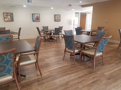 A dining room with several square wooden tables and chairs with blue patterned cushions. The room has wood flooring, beige and white walls, recessed ceiling lights, and framed artwork on the walls.
